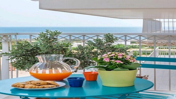 Un balcon avec une table préparée pour le petit-déjeuner, comprenant un pichet de jus d’orange, une assiette de biscuits et une plante en pot aux fleurs roses.