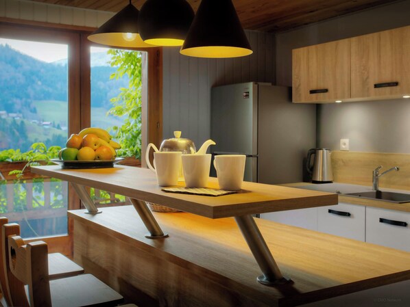 A modern kitchen with a wooden countertop, a sink, and a refrigerator. There is a window with a view of greenery and mountains.