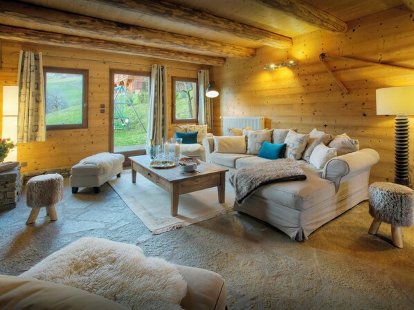 Ein gemütliches Blockhaus-Interieur mit Sofa, Hocker und Couchtisch, Holzwänden und Blick ins Freie.