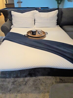 Un lit bien fait, avec un plateau sur lequel reposent deux tasses et une soucoupe, une couverture sombre et un oreiller blanc.