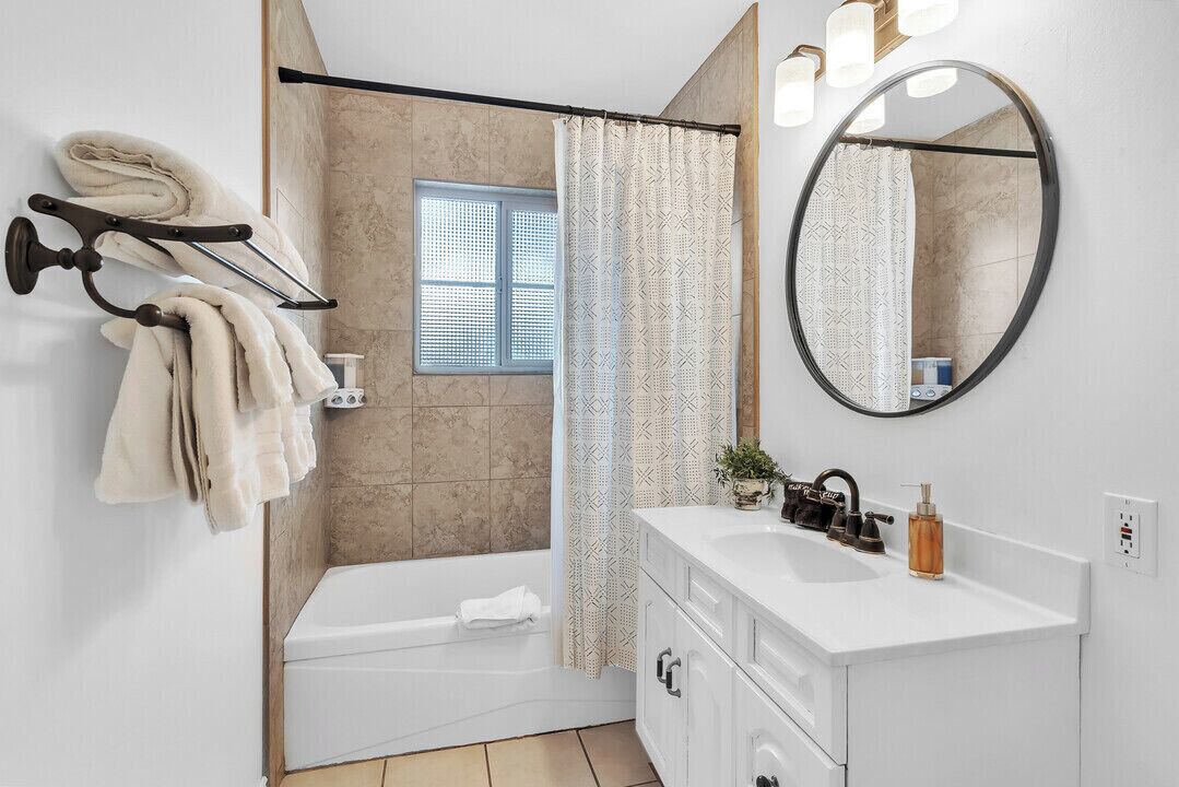 A modern bathroom with a bathtub, a shower curtain, a towel rack, a sink, a mirror, and a window.