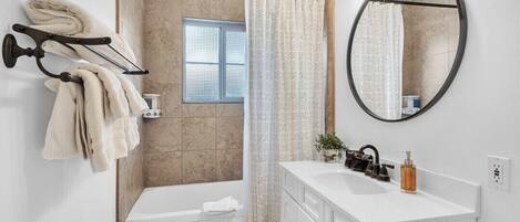 A modern bathroom with a bathtub, a shower curtain, a towel rack, a sink, a mirror, and a window.