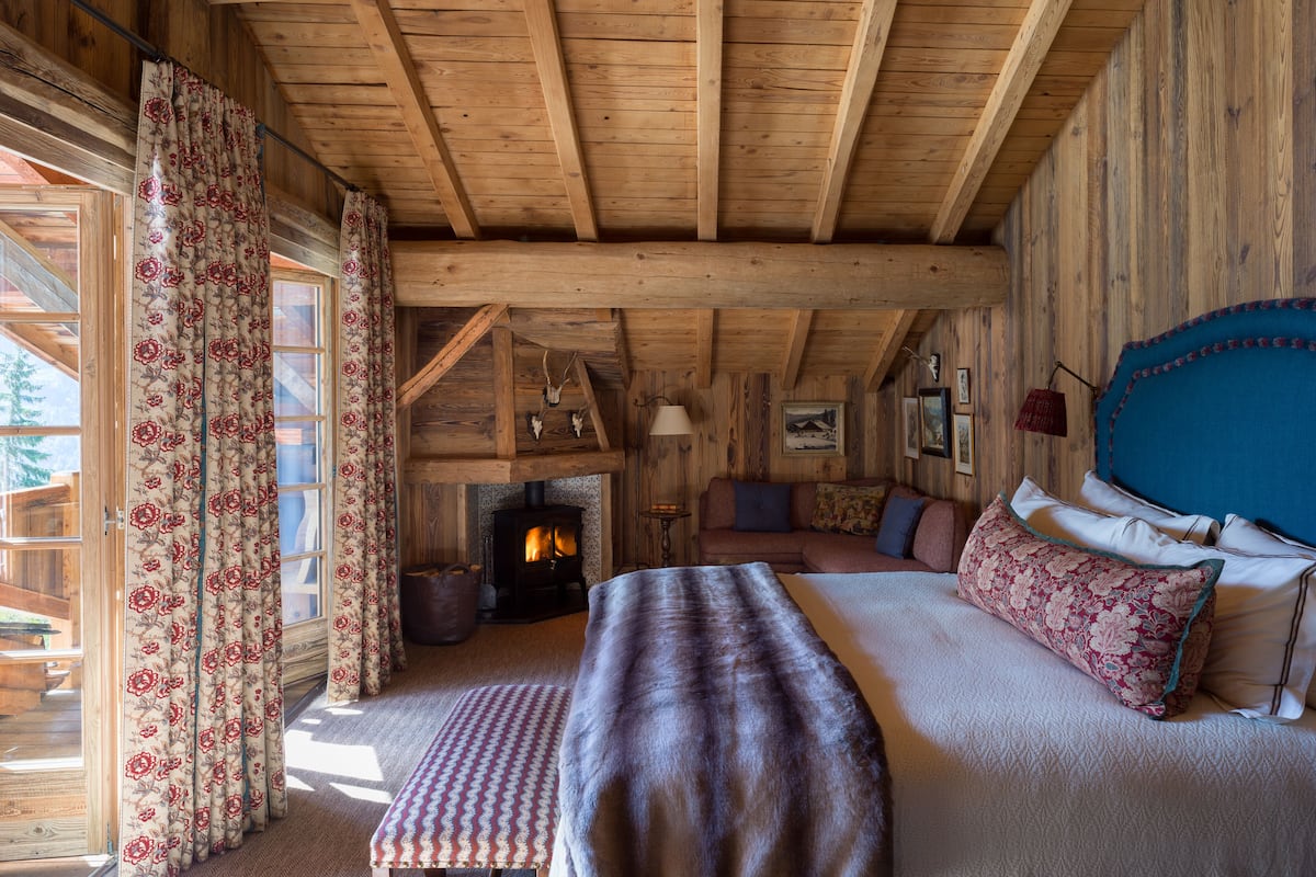 Un interior de cabaña acogedor con techo de madera, chimenea, cama con ropa de cama floral y un ventanal con cortinas.