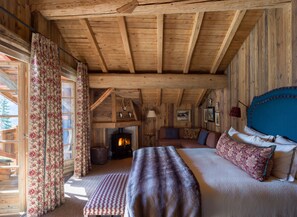 L’intérieur d’un chalet accueillant, avec un plafond en bois, une cheminée, un lit recouvert d’une literie à motifs floraux et une grande fenêtre agrémentée de rideaux.