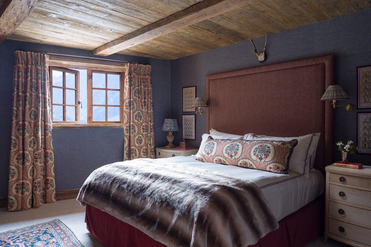 Un dormitorio con techo de madera, una cama grande con ropa de cama estampada, una mesita de noche con lámpara y una ventana con cortinas.