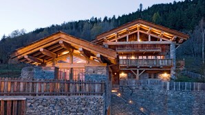 Ein mehrstöckiges Blockhaus mit Steinmauern und hölzernen Balkonen, eingebettet in einen bewaldeten Hügelhang.