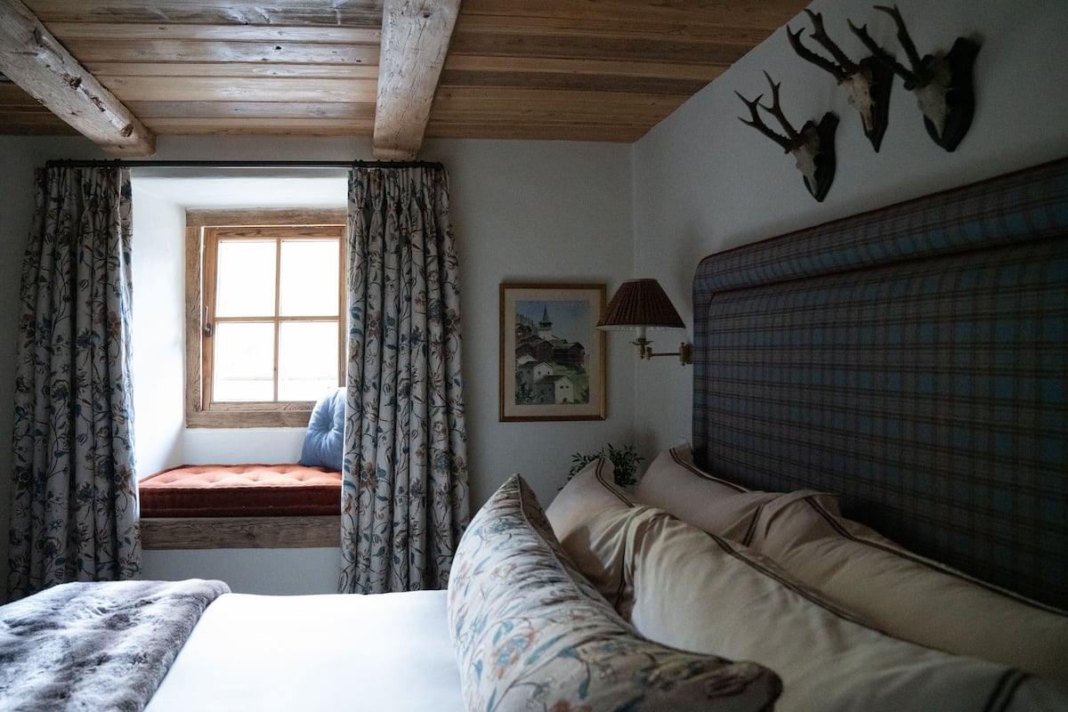 Un dormitorio con techo de madera, ventana con cortinas florales, cama con ropa de cama estampada, cabecera con diseño escocés y cabezas de animales montadas en la pared.