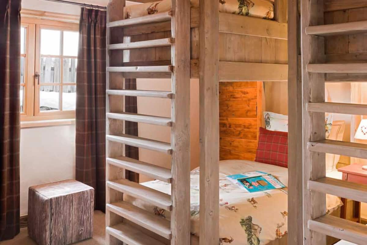 Una habitación rústica con literas, vigas de madera, una ventana con cortinas y un taburete pequeño.
