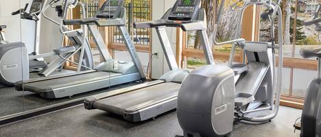 A gym with treadmills and elliptical machines, large windows, and a view of trees and buildings outside.