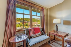 Une pièce confortable donnant sur la ville, avec un canapé agrémenté d’un plaid rouge, une petite table avec une lampe et une fenêtre avec des rideaux.