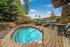 Un bain à remous avec un mur en pierre et une terrasse en bois, entouré d’une clôture en pierre et d’arbres.