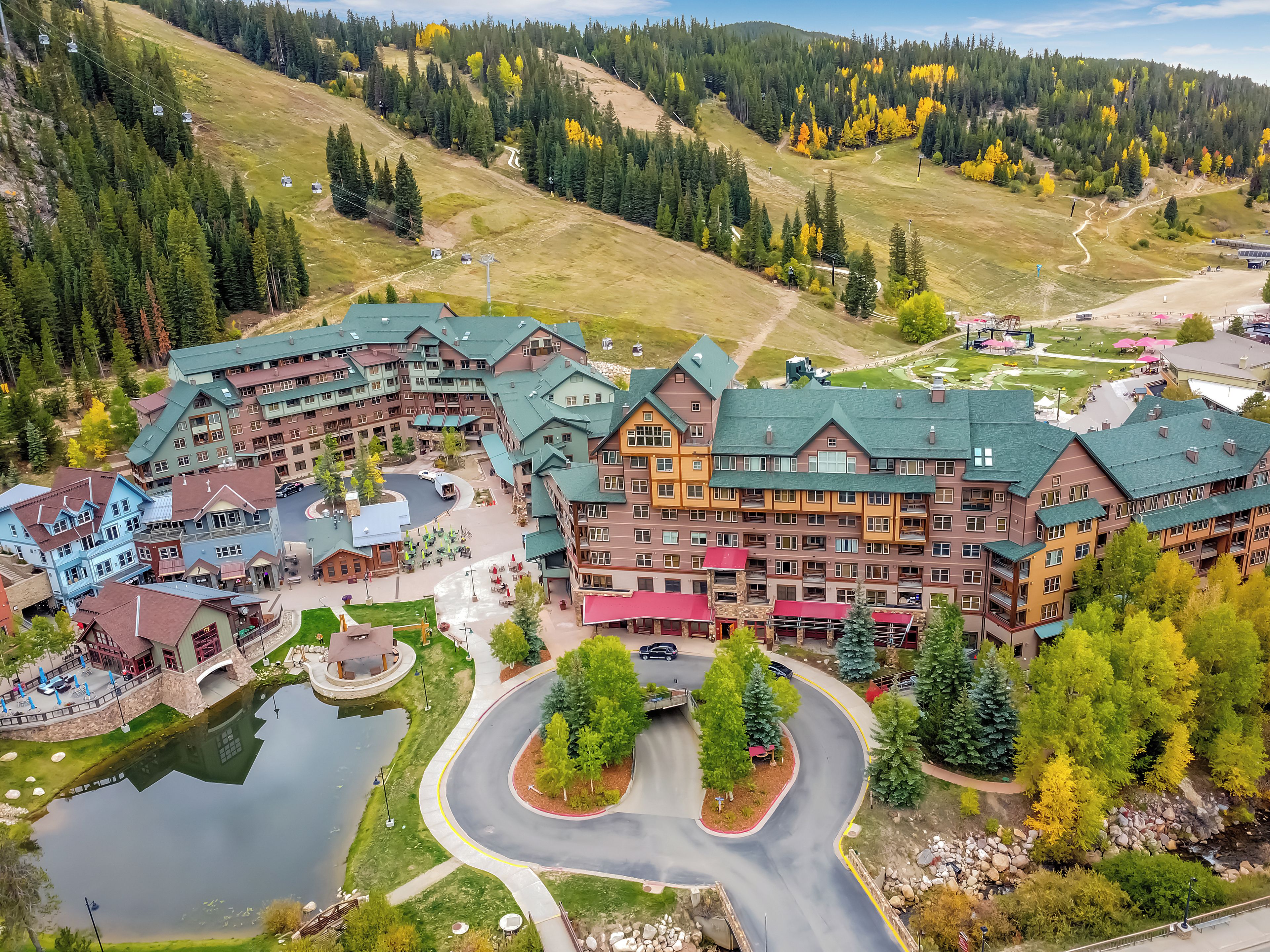A resort with a central building surrounded by a lake, a ski lift, and a mountainous backdrop.