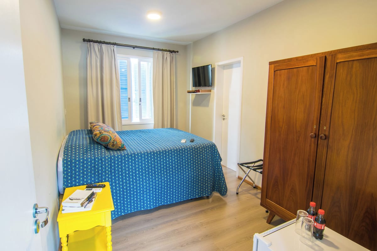 Ein Schlafzimmer mit einem Bett, einem Holzschrank, einem an der Wand montierten Fernseher und einem Fenster mit Vorhängen.