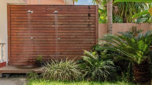 Une clôture en bois agrémentée d’un élément décoratif avec de l’eau, entourée d’une végétation luxuriante et bordée d’un chemin en béton.