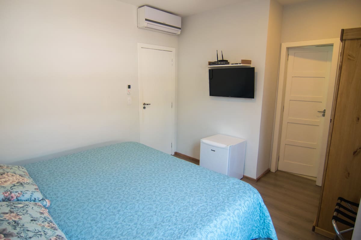 Ein Schlafzimmer mit einem Bett, einem weißen Kleiderschrank, einer kleinen weißen Kommode, einem an der Wand montierten Fernseher und einer Klimaanlage.
