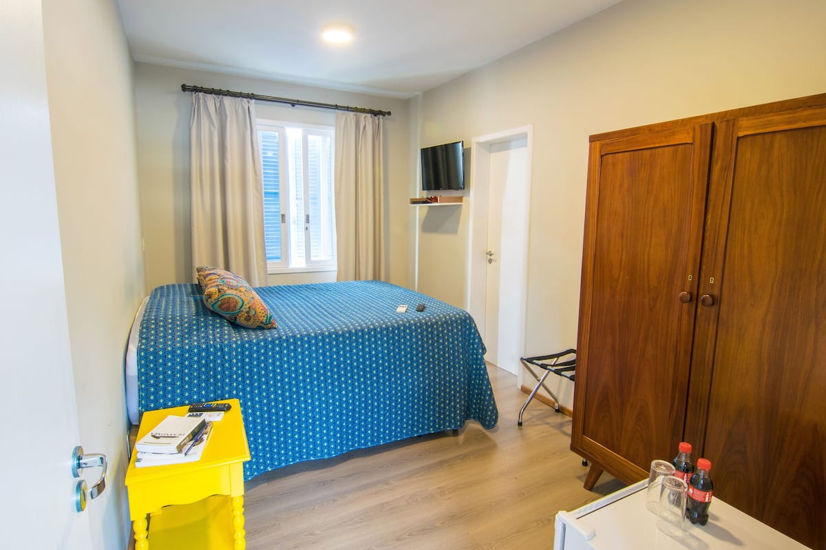 Ein Schlafzimmer mit einem Bett, einem Holzschrank, einem an der Wand montierten Fernseher und einem Fenster mit Vorhängen.