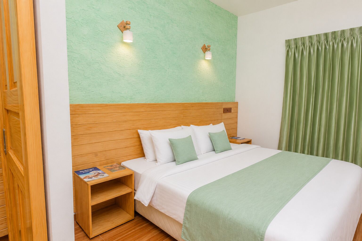 Habitación de hotel con cabecera de madera, pared verde, cama con ropa blanca y almohadas verdes, mesita de noche con un libro y cortina verde.
