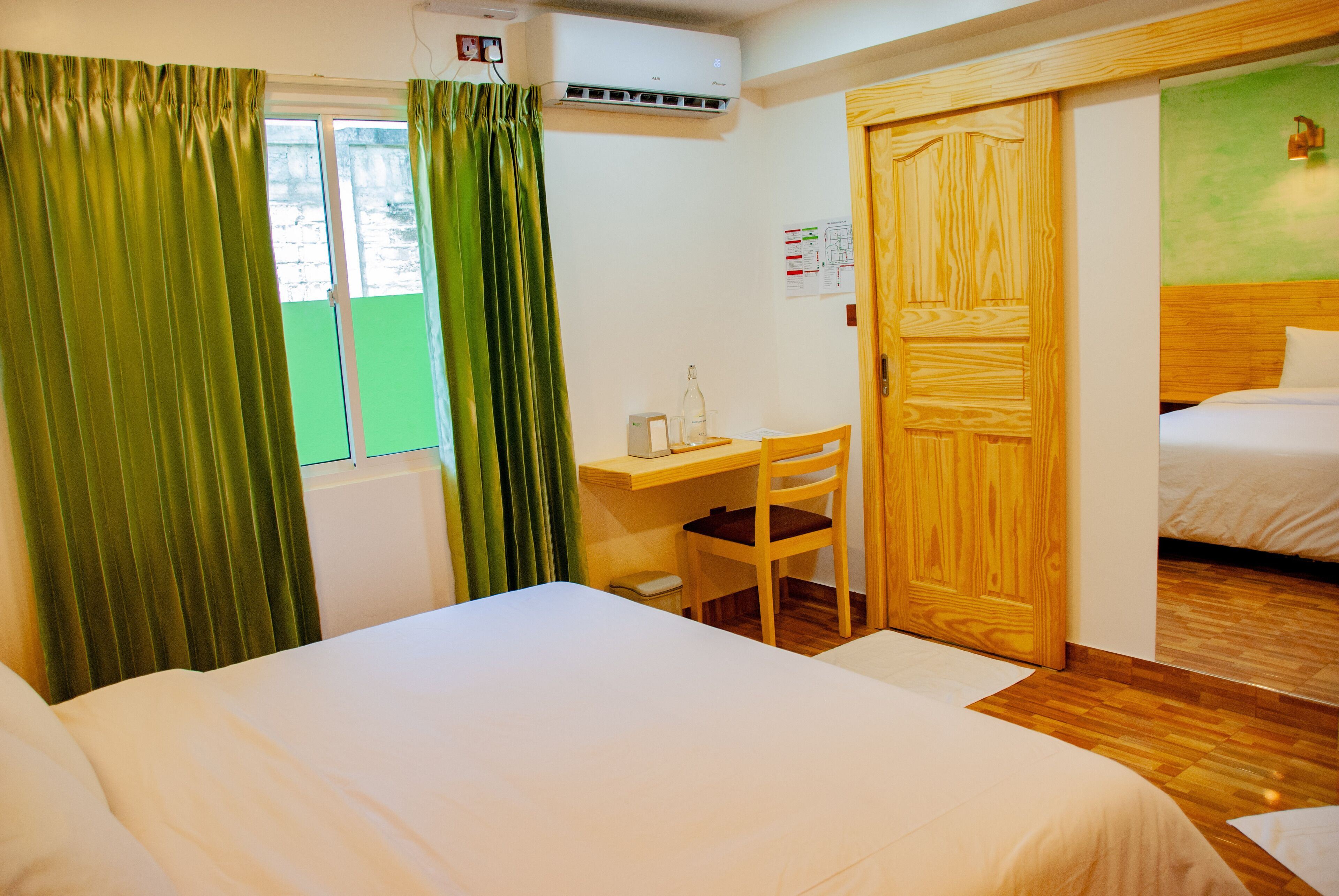 A hotel room with a bed, a desk with a chair, a wooden wardrobe, and a window with curtains.