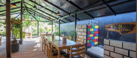 An indoor dining area with wooden tables and chairs, a glass wall with a colorful brick pattern, and a sign reading "KIDS CORNER".