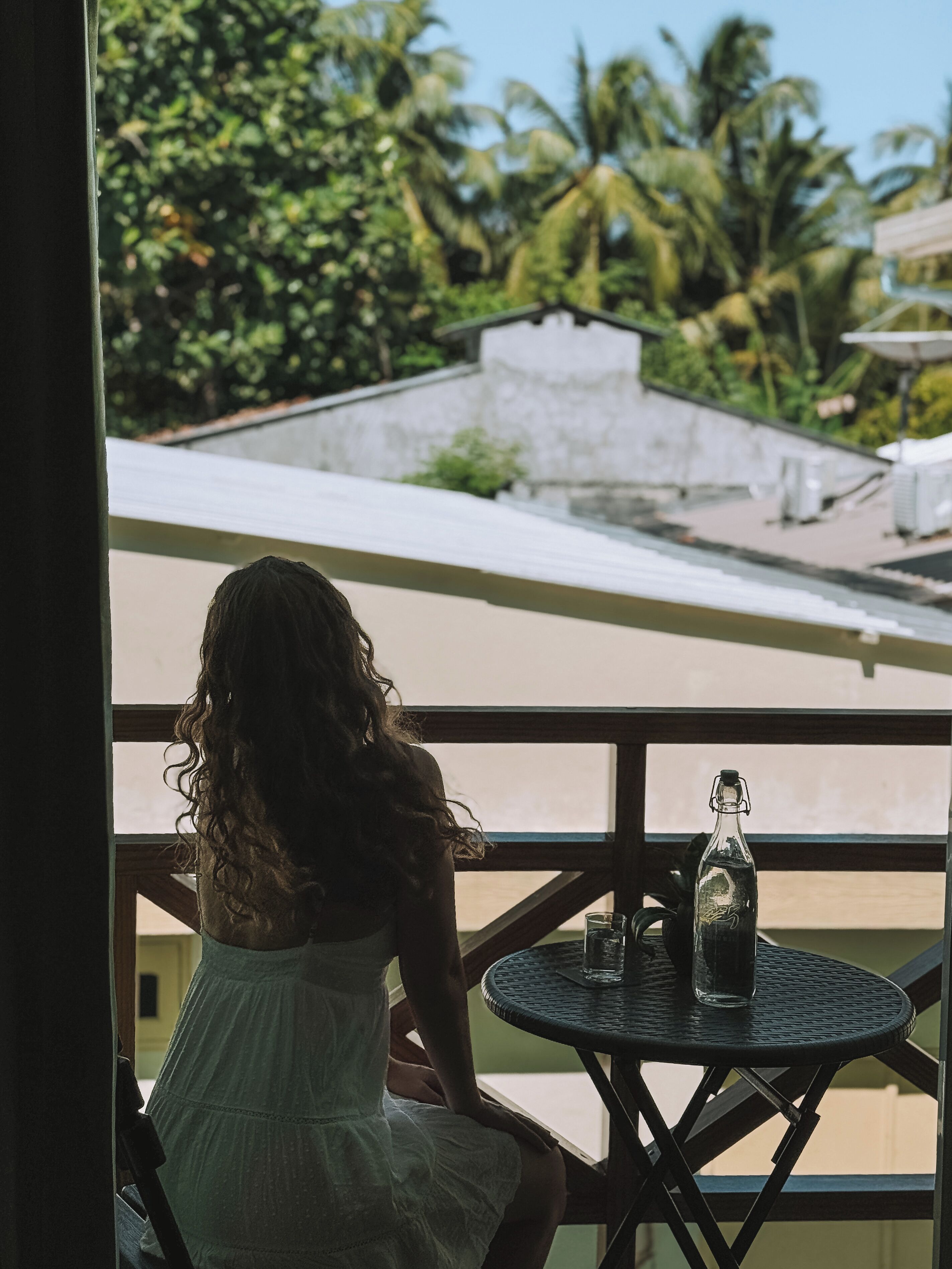 Una mujer con vestido blanco sentada a una mesa redonda con una botella y un vaso, con vista a un balcón que ofrece vistas de vegetación y edificios.