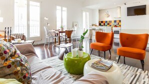 Un salon moderne comprenant un canapé, deux fauteuils orange, un pouf vert et un coin repas avec une table et des chaises.