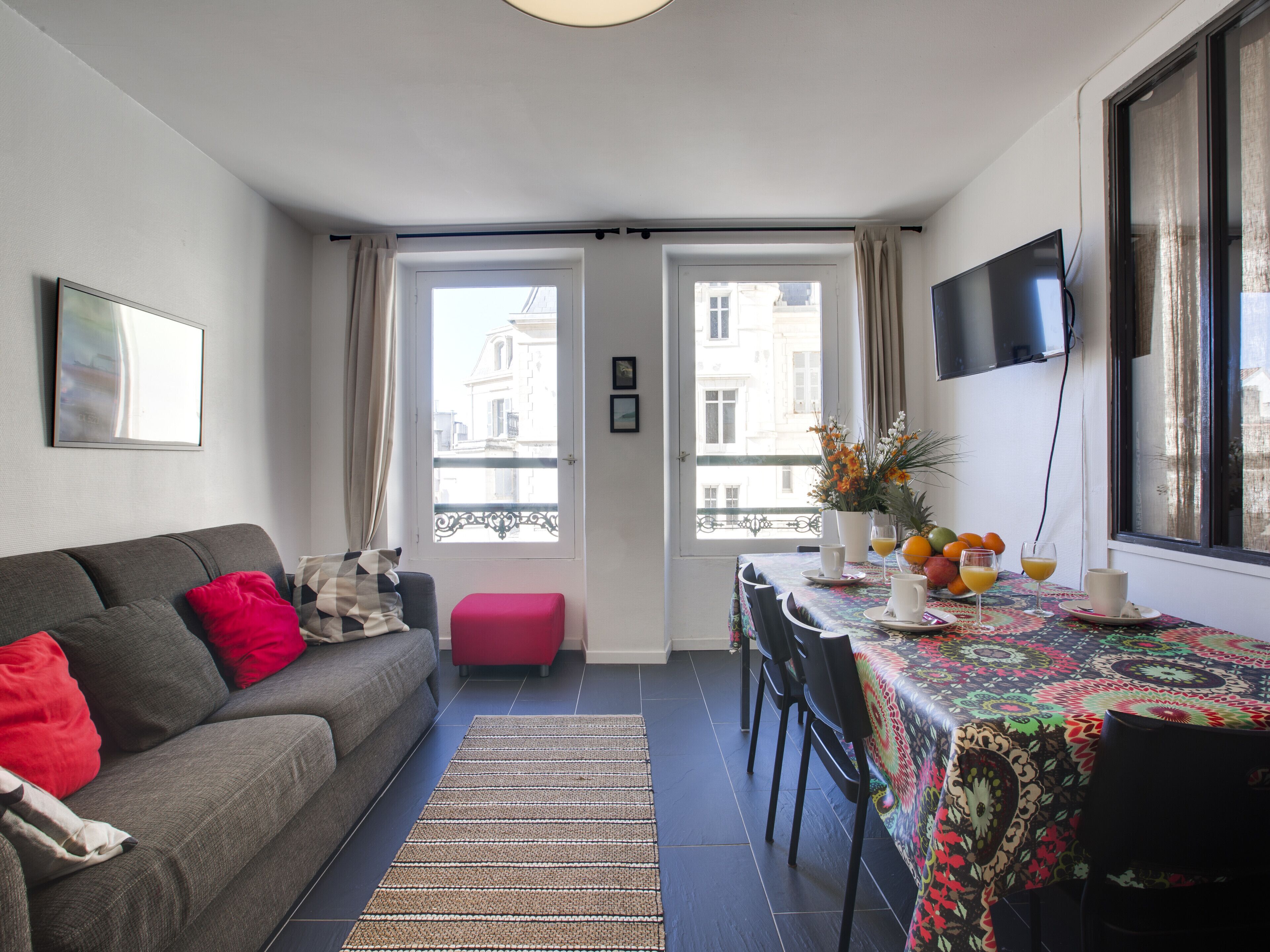 A modern living room with a sofa, a dining table with chairs, a TV, and a balcony with a view of buildings.