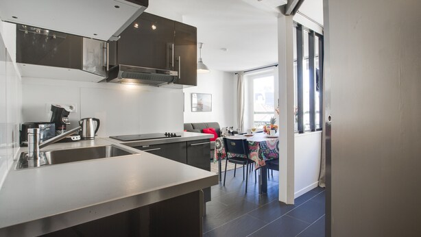 A modern kitchen with stainless steel appliances, a dining area with a table and chairs, and a view of the outside through large windows.