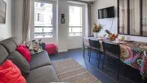 A modern living room with a grey sofa, a dining table with chairs, and a large window with curtains.