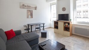A modern living room with a grey sofa, a small ottoman, a coffee table, a TV on a wooden stand, and a dining area with a table and chairs.
