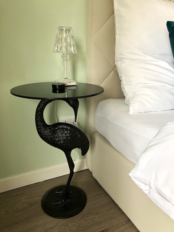 Een hoek van een kamer met een ronde glazen tafel, een zwart decoratief voetstuk, een lamp en een bed met witte beddengoed.