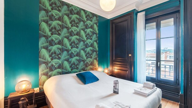 A hotel room with a large bed, a nightstand with a lamp, a window with a view, and a tropical wallpaper. - Appartement Majorel - Welkeys (Lyon)