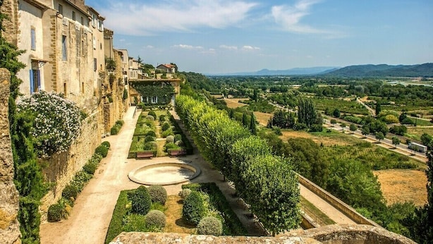 A historic building with a garden pathway leading to a circular fountain, surrounded by lush greenery and a distant view of rolling hills. - Comfortable 3 bedroom mobile home with air conditioning in Provence - Swimming pool and private lake (La Roque-d'Anthéron)