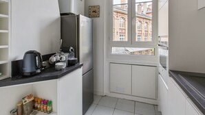Une cuisine compacte équipée d’un réfrigérateur, d’une bouilloire et d’une théière sur le plan de travail. Une fenêtre offre une vue sur les bâtiments environnants.