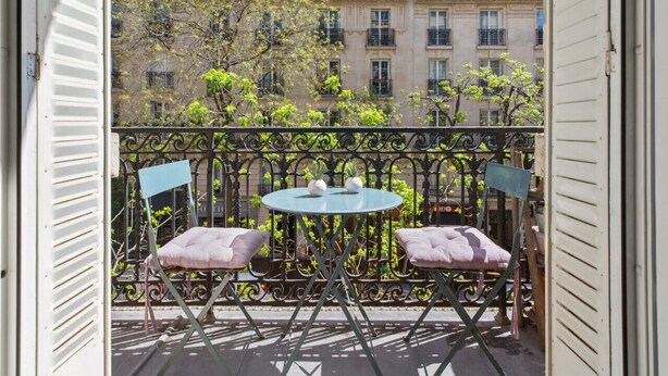 Un balcon avec une table ronde et deux chaises, donnant sur une rangée d’immeubles dotés de balcons.