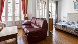 Une pièce avec un plancher en bois, un canapé en cuir, un lit recouvert d’une courtepointe à motifs et une table avec une lampe.