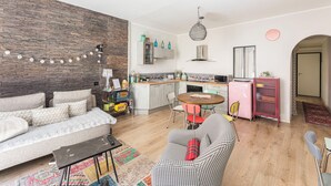A modern living room with a sofa, a houndstooth armchair, a dining table with chairs, a kitchen area with cabinets and appliances, and a wooden floor.