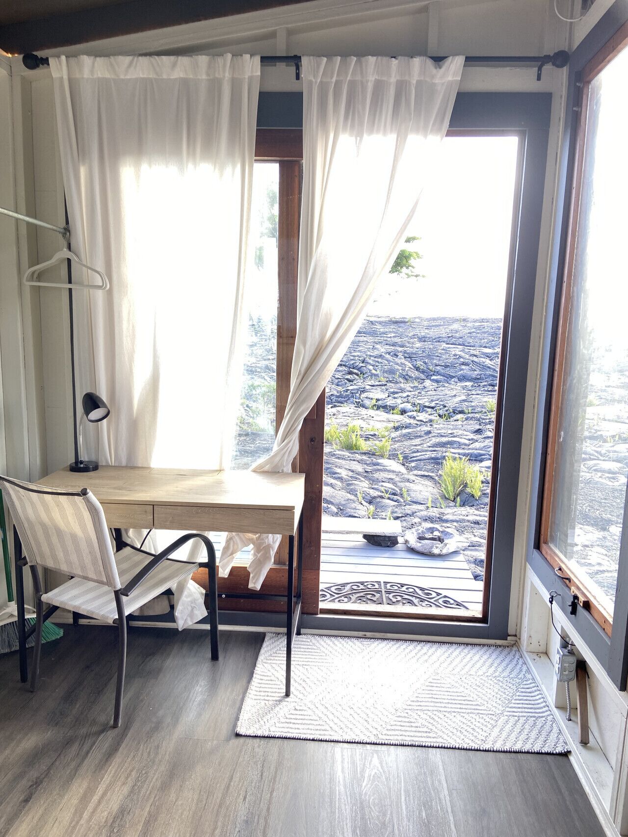 A room with a wooden desk, a chair, a hanging lamp, and a view of a rocky landscape through a large window with sheer curtains.