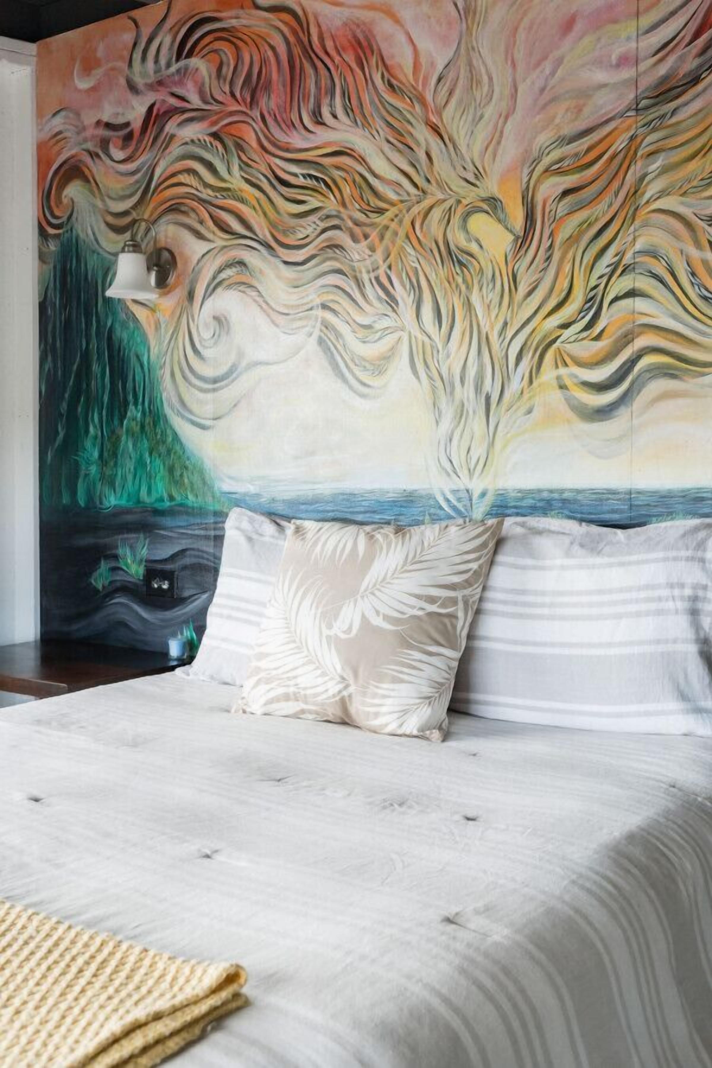 A bed with a textured white bedspread, two pillows with leaf patterns, and a colorful abstract mural on the wall behind.