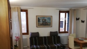 Una sala de estar con sofá, una mesa de comedor con sillas, un cuadro en la pared y un ventanal con cortinas.