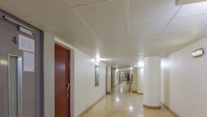 A long, narrow hallway with a tiled floor, white walls, and a ceiling with a perforated pattern. There are doors on the left, one of which has a number sign.