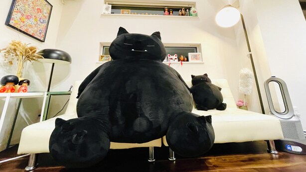 A room with a large black cat sculpture on a white chair, a small black cat on the floor, and a shelf with decorative items.
