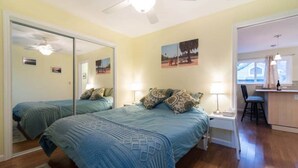 A bedroom with two beds, a ceiling fan, a bedside table with a lamp, and a small kitchenette area. - Light and bright family-friendly cottage (Portland)