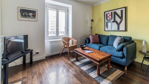 A living room with a TV, wicker chair, blue sofa, and a framed picture on the wall. - Appartement Tromba - Welkeys (Lyon)