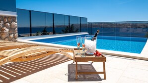 Uma área à beira da piscina com espreguiçadeira de madeira, mesa com taças de champanhe e gelo, e vista para o mar.