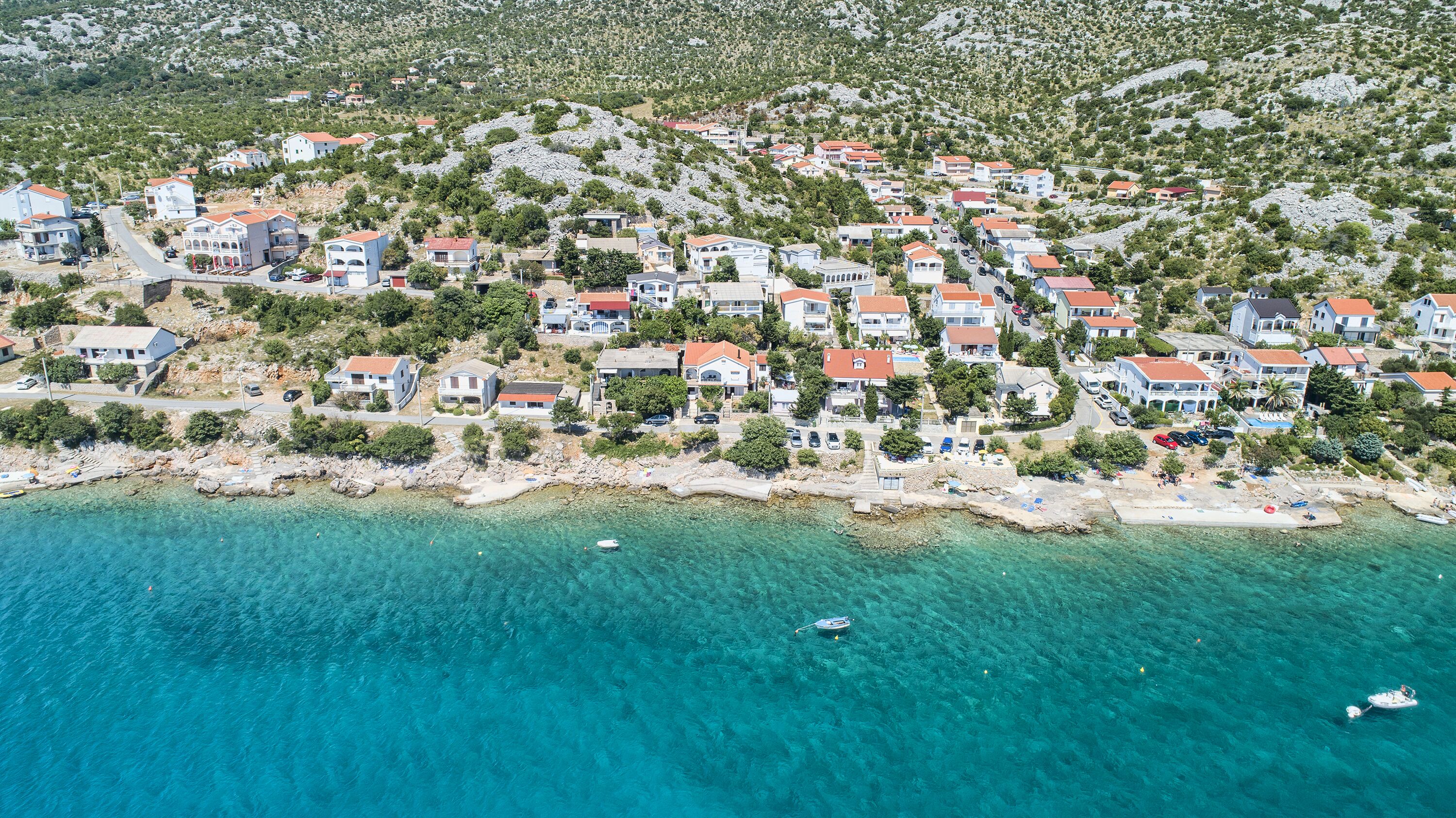 An aerial view of a coastal residential area with houses, clear turquoise waters, and a rocky hillside.