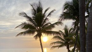 Duas poltronas azuis em uma praia arenosa, com palmeiras e vista para o oceano ao pôr do sol.