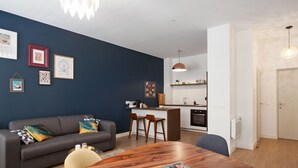 Eine moderne Wohnung mit dunkelblauer Akzentwand, einem Holzesstisch, einem grauen Sofa mit gemusterten Kissen und einer Küchenzeile mit weißen Schränken und einer hängenden Pendelleuchte.