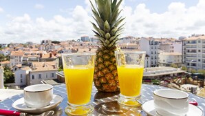 Ein Tisch mit zwei Orangensaftgläsern, einer Ananas und zwei kleinen weißen Tassen auf einem Dach mit Blick auf eine Stadtlandschaft.