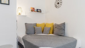 Une chambre à coucher avec un lit, des oreillers, une étagère remplie de livres, une horloge murale et une petite table.