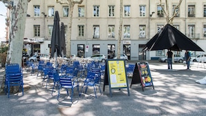 Terrasse extérieure avec des chaises et des parasols bleus, un panneau indicateur et, au fond, un bâtiment doté de nombreuses fenêtres.